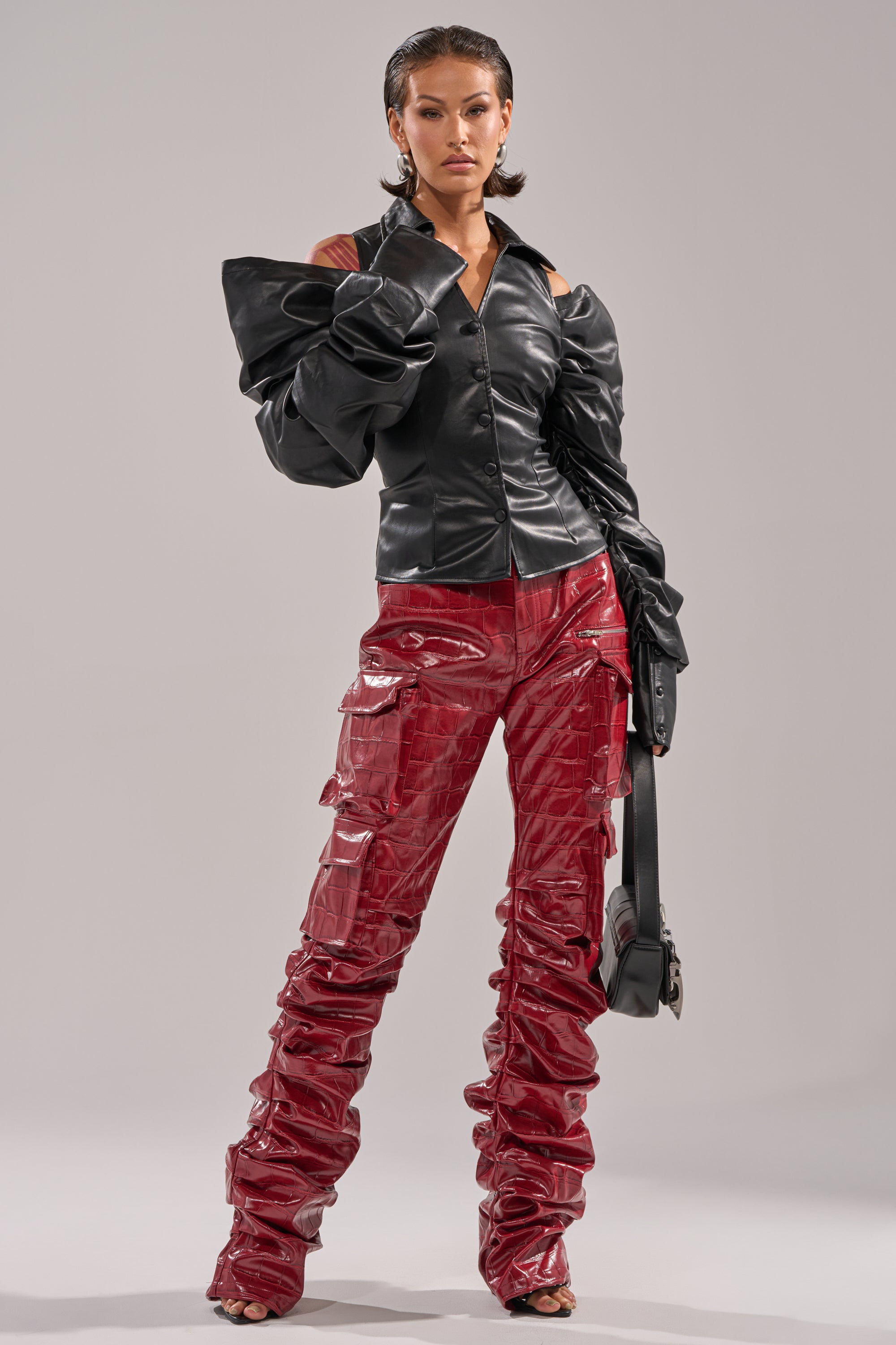 A woman models the CRAZY IN LOVE BLACK FAUX LEATHER BUTTON DOWN BLOUSE with dramatic ruched sleeves, paired with shiny red pants and a black shoulder bag, standing confidently against a plain background. 6