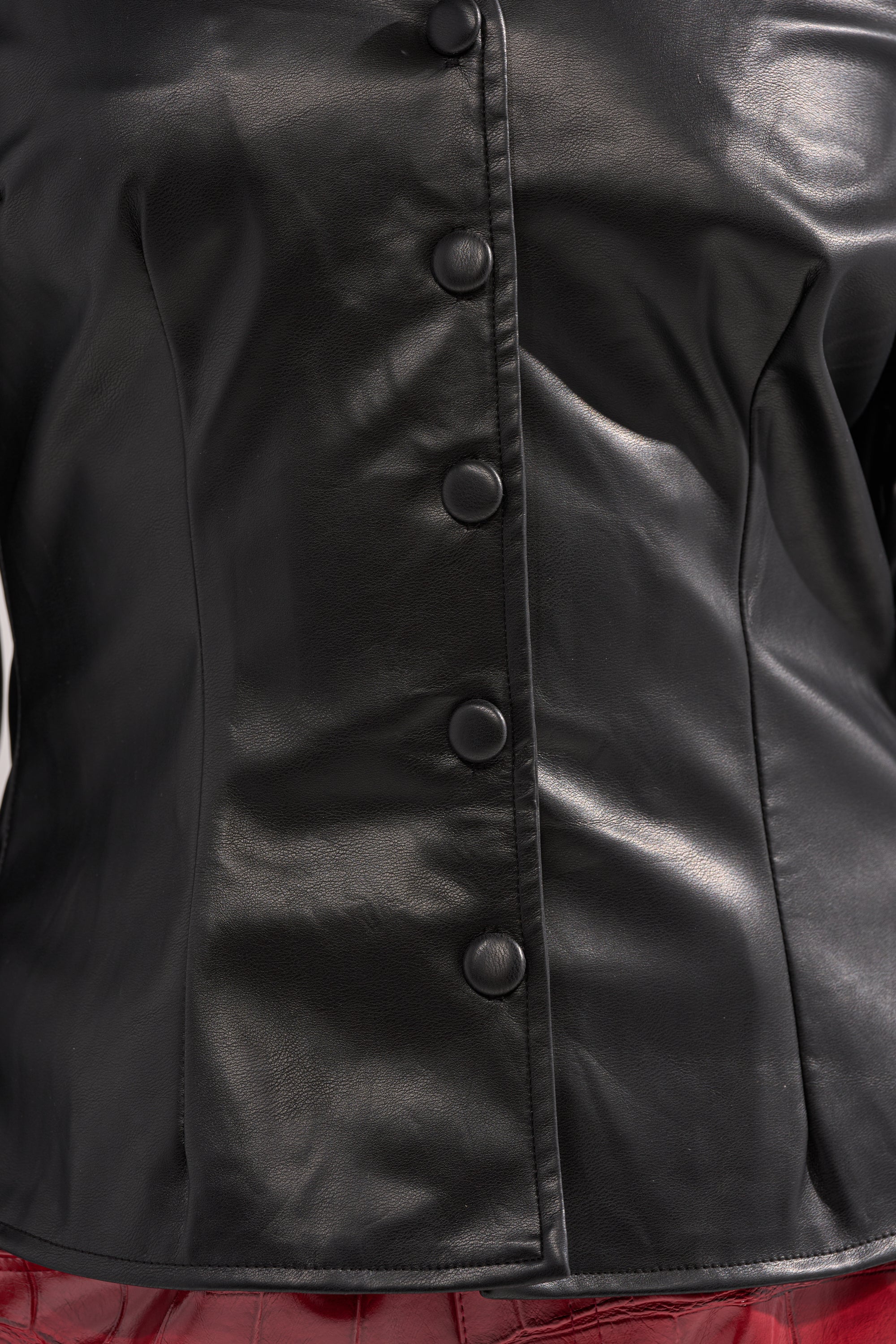 Close-up of a torso wearing the CRAZY IN LOVE BLACK FAUX LEATHER BUTTON DOWN BLOUSE with large round black buttons and ruched sleeves, paired with glossy red bottoms. 7