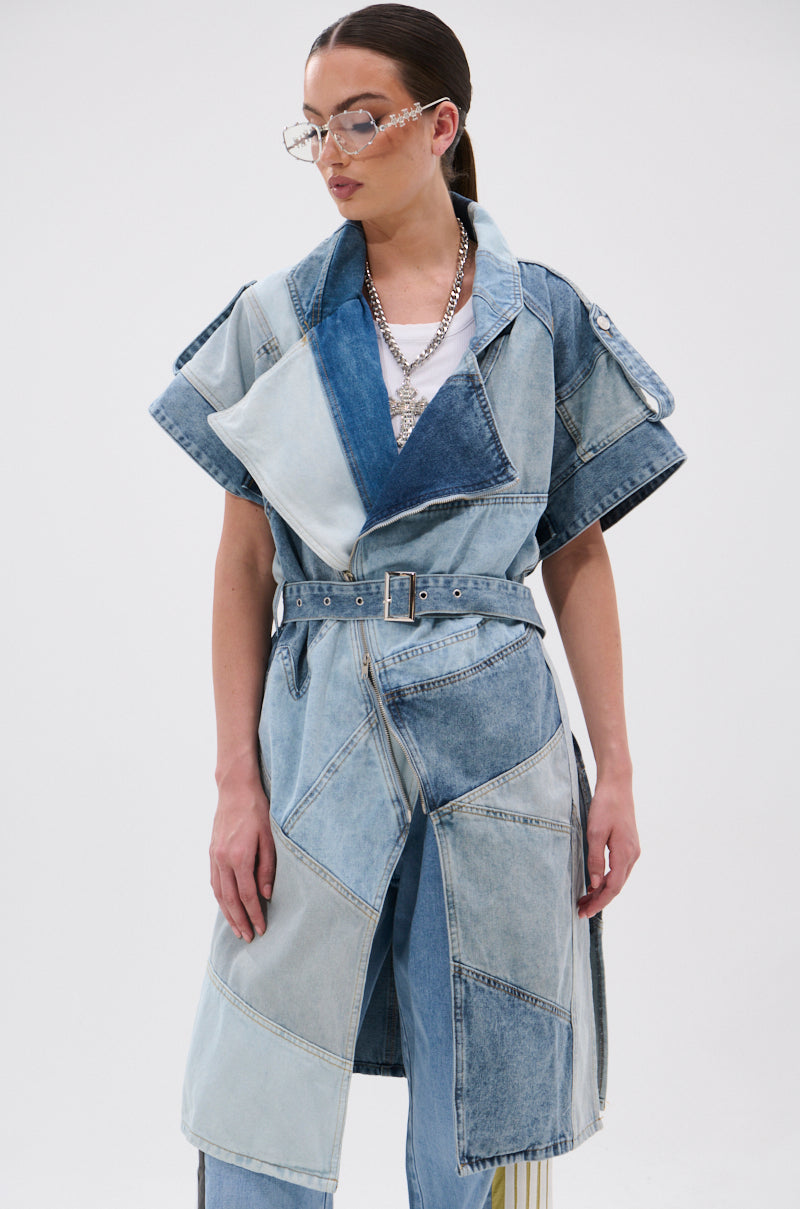 A person stands against a plain white background wearing the ESTELLE PATCHWORK DENIM VEST over a white shirt and blue jeans, styled with layered necklaces, clear glasses, and slicked-back hair.