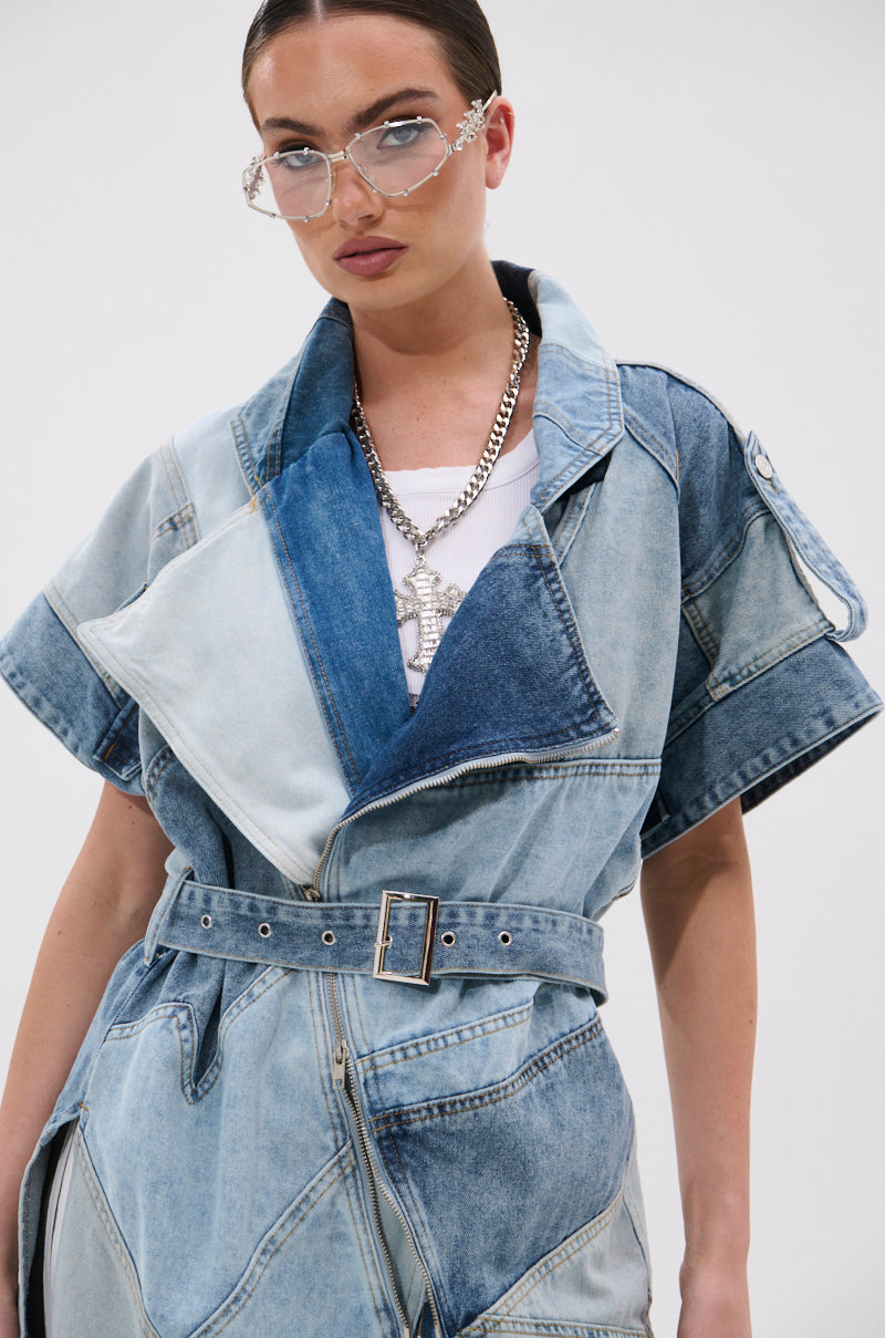 A woman makes a streetwise statement in the ESTELLE PATCHWORK DENIM VEST with a belted waist, silver chain necklace, clear-framed glasses, and slicked-back hair as she poses confidently against a plain white background.