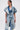 A person stands against a plain white background wearing the ESTELLE PATCHWORK DENIM VEST over a white shirt and blue jeans, styled with layered necklaces, clear glasses, and slicked-back hair.