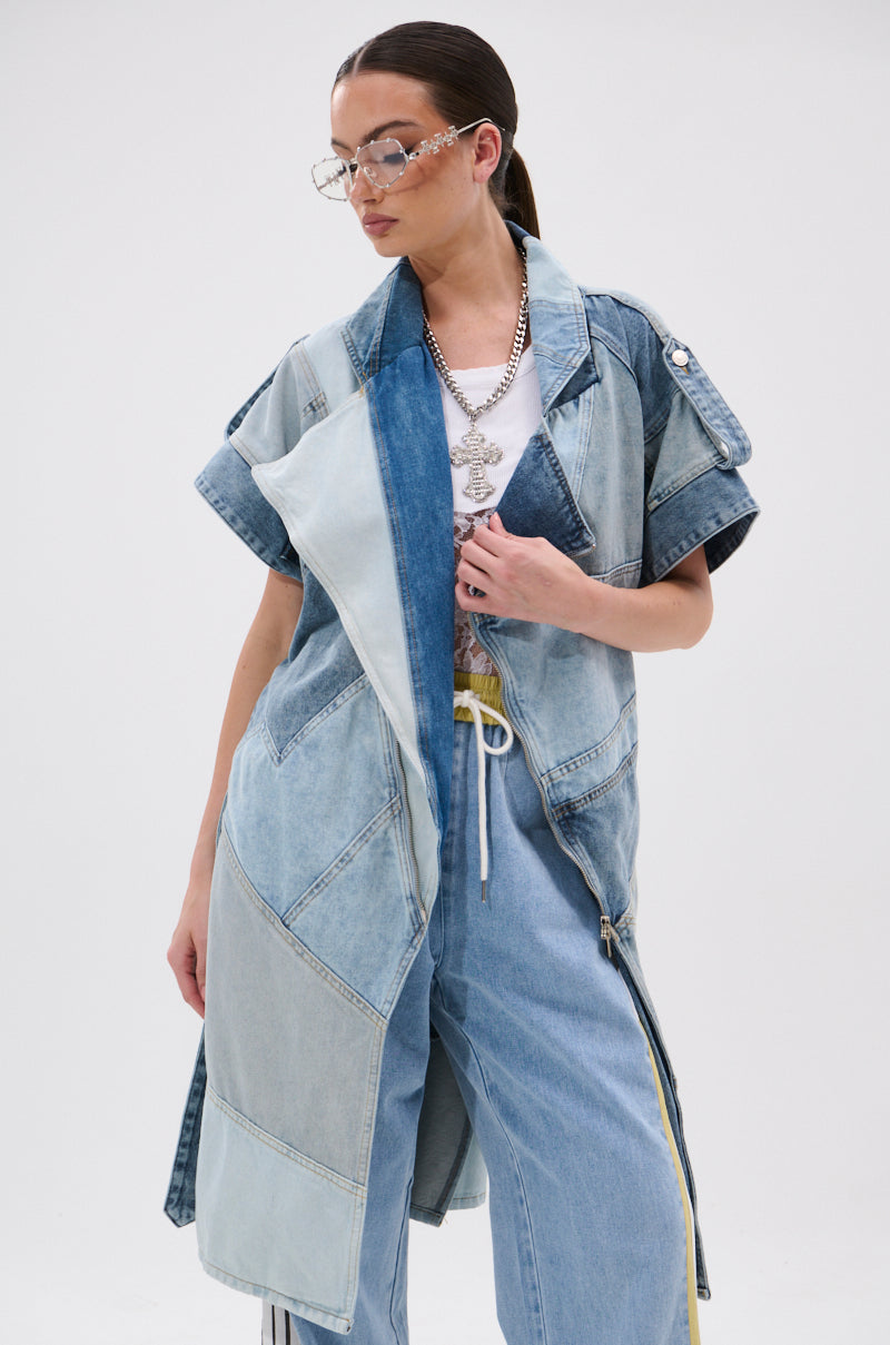 A person wearing the ESTELLE PATCHWORK DENIM VEST over wide-leg jeans, a white t-shirt, layered necklaces, and large sunglasses stands against a plain white background.