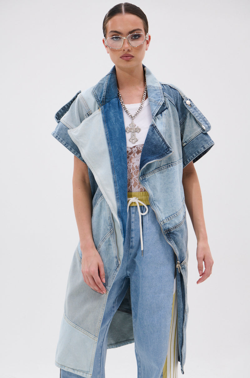 Wearing the ESTELLE PATCHWORK DENIM VEST over a white lace top and blue jeans, a woman with slicked-back hair, glasses, and a chunky pendant necklace poses confidently against a plain white background.