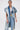 Wearing the ESTELLE PATCHWORK DENIM VEST over a white lace top and blue jeans, a woman with slicked-back hair, glasses, and a chunky pendant necklace poses confidently against a plain white background.