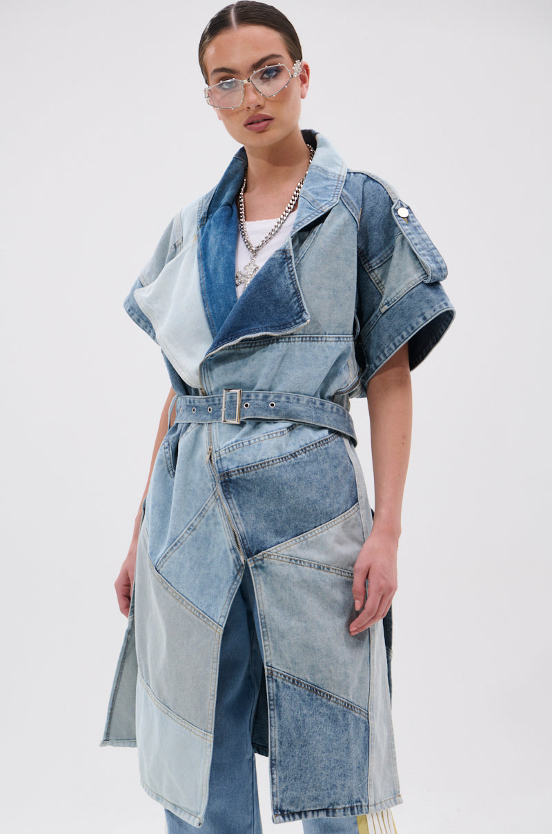A woman in transparent glasses and layered necklaces models the ESTELLE PATCHWORK DENIM VEST—a belted, trench-style, short-sleeve denim vest—over blue jeans against a plain white background.
