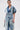 A woman in transparent glasses and layered necklaces models the ESTELLE PATCHWORK DENIM VEST—a belted, trench-style, short-sleeve denim vest—over blue jeans against a plain white background.