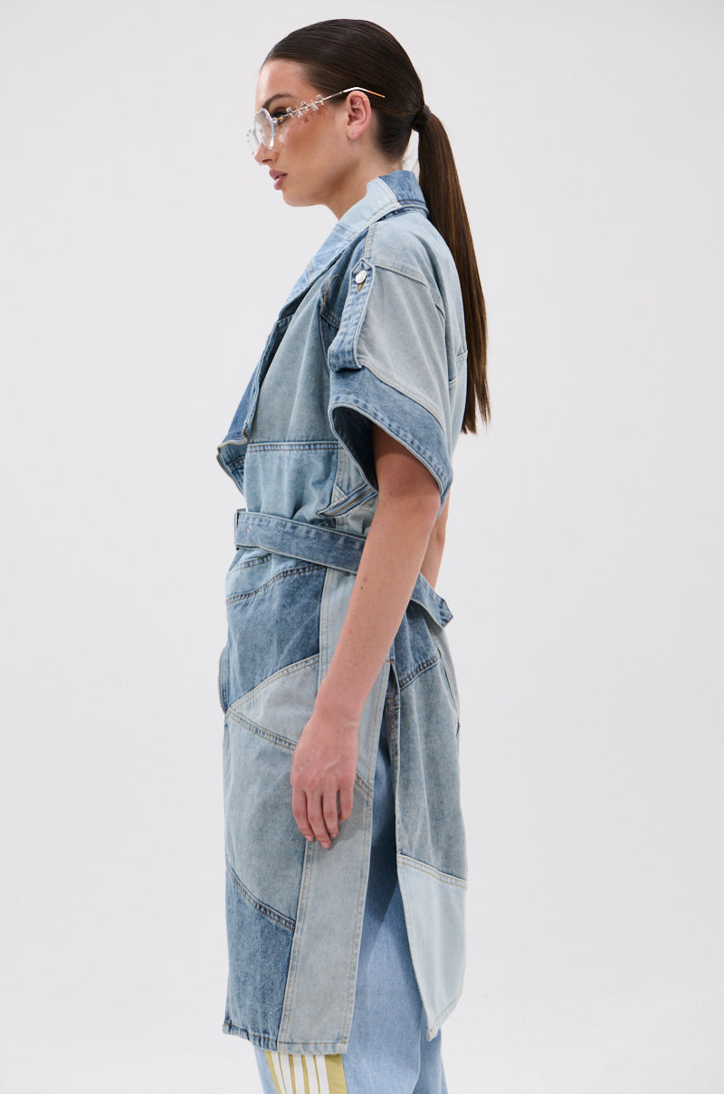 A person with long dark hair in a ponytail wears clear glasses and the ESTELLE PATCHWORK DENIM VEST over light blue pants, standing in profile against a plain white background.