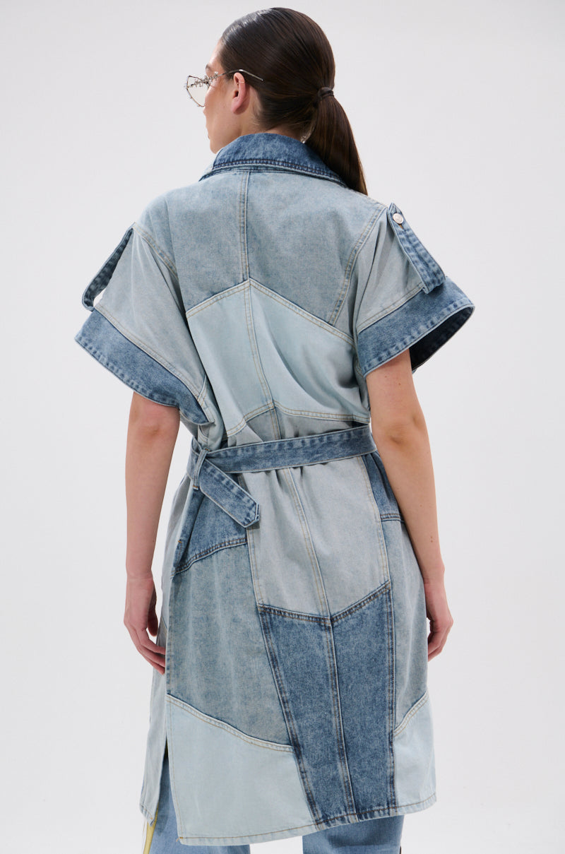 A woman with tied-back brown hair and glasses faces away, wearing the ESTELLE PATCHWORK DENIM VEST—a trench-style denim vest featuring visible patchwork seams and a belt—against a plain white background.
