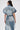 A woman with tied-back brown hair and glasses faces away, wearing the ESTELLE PATCHWORK DENIM VEST—a trench-style denim vest featuring visible patchwork seams and a belt—against a plain white background.