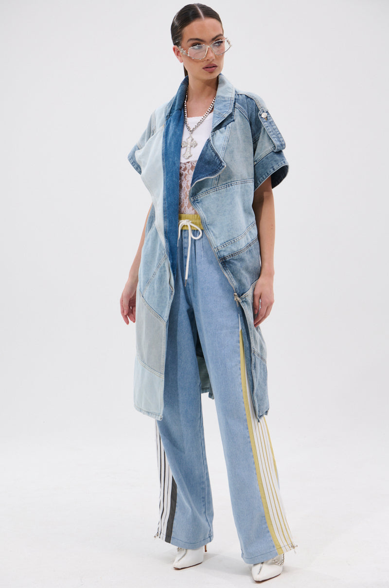 A woman stands confidently in the ESTELLE PATCHWORK DENIM VEST over a white top, paired with light blue jeans featuring yellow and black stripes, white pointed heels, clear glasses, and layered necklaces against a white background.
