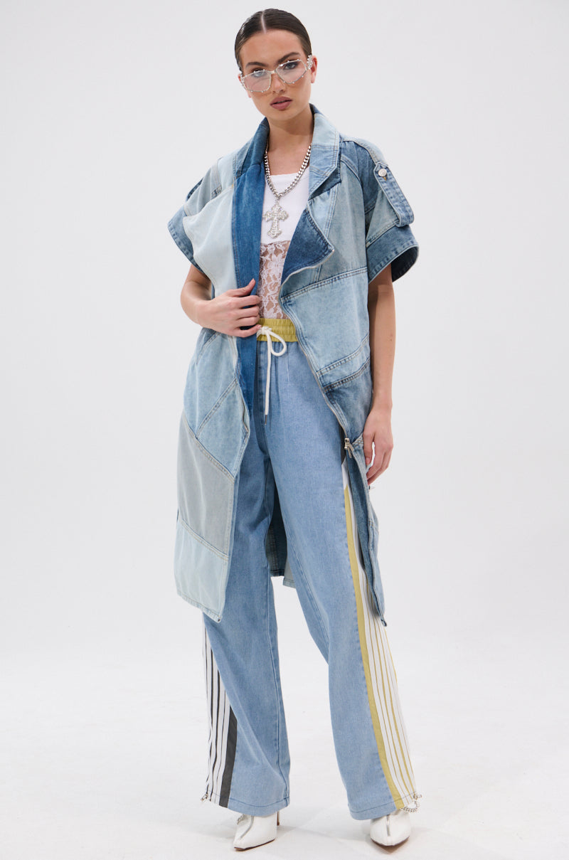 A woman stands confidently in the ESTELLE PATCHWORK DENIM VEST and wide-leg yellow and white striped jeans, with a lace top, white pointed shoes, glasses, and layered necklaces against a plain white background.