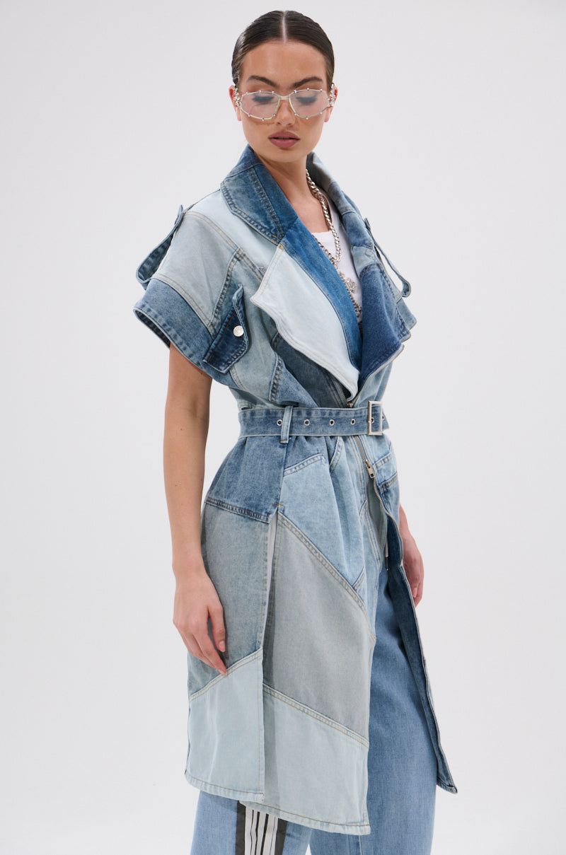 Wearing the ESTELLE PATCHWORK DENIM VEST with a belt over blue jeans and a blue top, this individual with slicked-back hair and clear glasses poses against a plain white background.