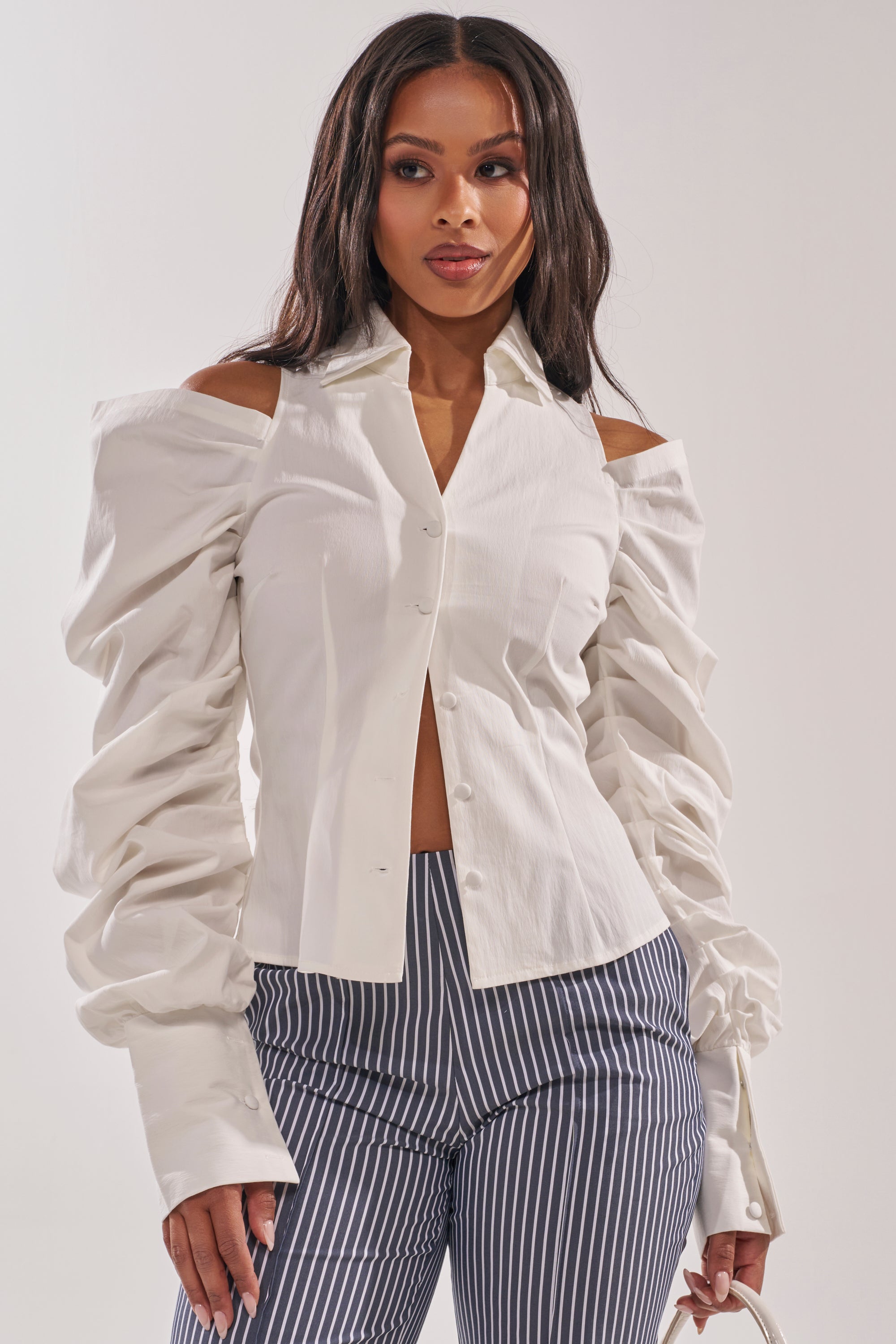 A woman stands confidently to the side, holding a small white handbag and wearing the CRAZY IN LOVE WHITE BUTTON DOWN BLOUSE with long, ruched sleeves and pinstriped gray pants. 0
