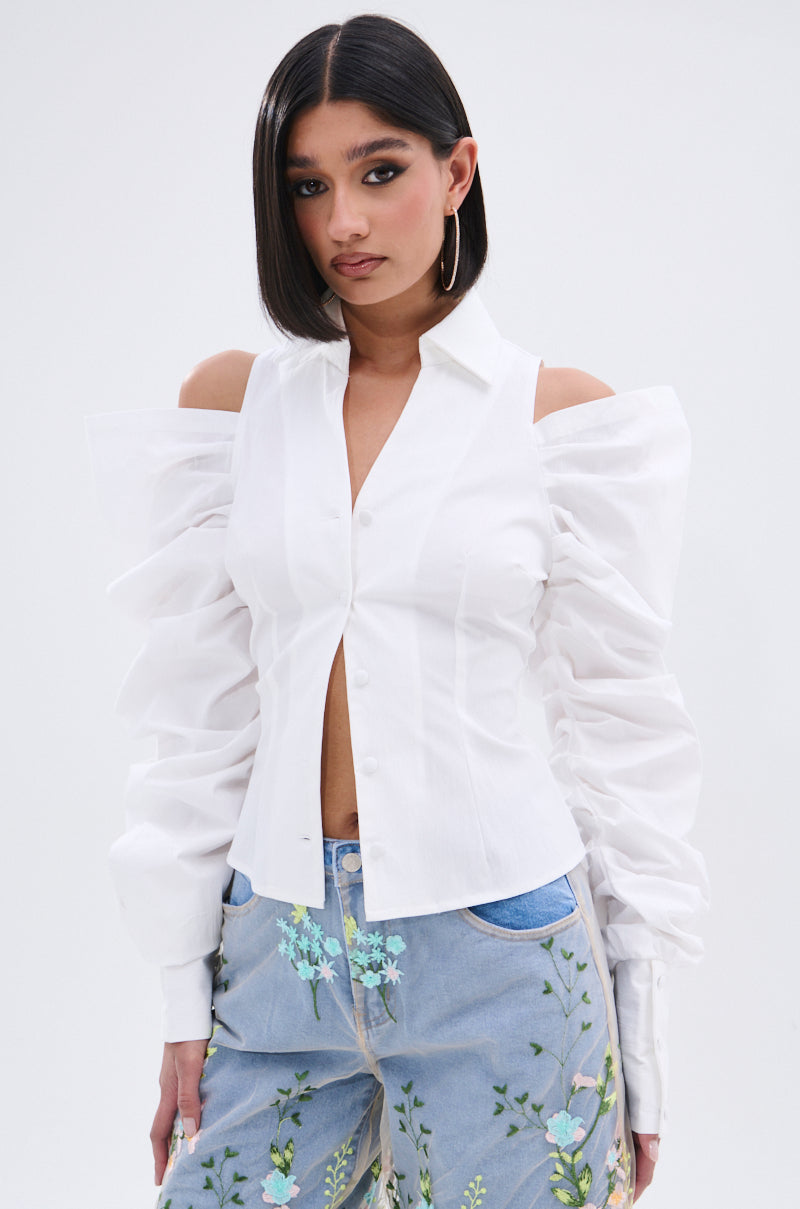 A woman with straight, shoulder-length dark hair wears the CRAZY IN LOVE BUTTON DOWN BLOUSE with ruched sleeves and blue jeans adorned with embroidered floral patterns, posing against a plain light background.