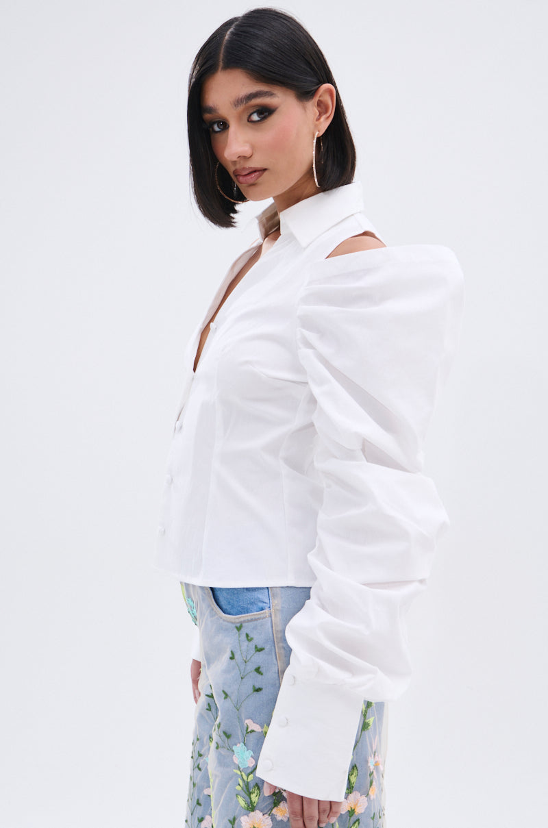 A woman with straight dark hair wears the CRAZY IN LOVE BUTTON DOWN BLOUSE, featuring white puffed statement sleeves and cold shoulder cut-outs, paired with light blue jeans with colorful floral embroidery, against a plain white background.