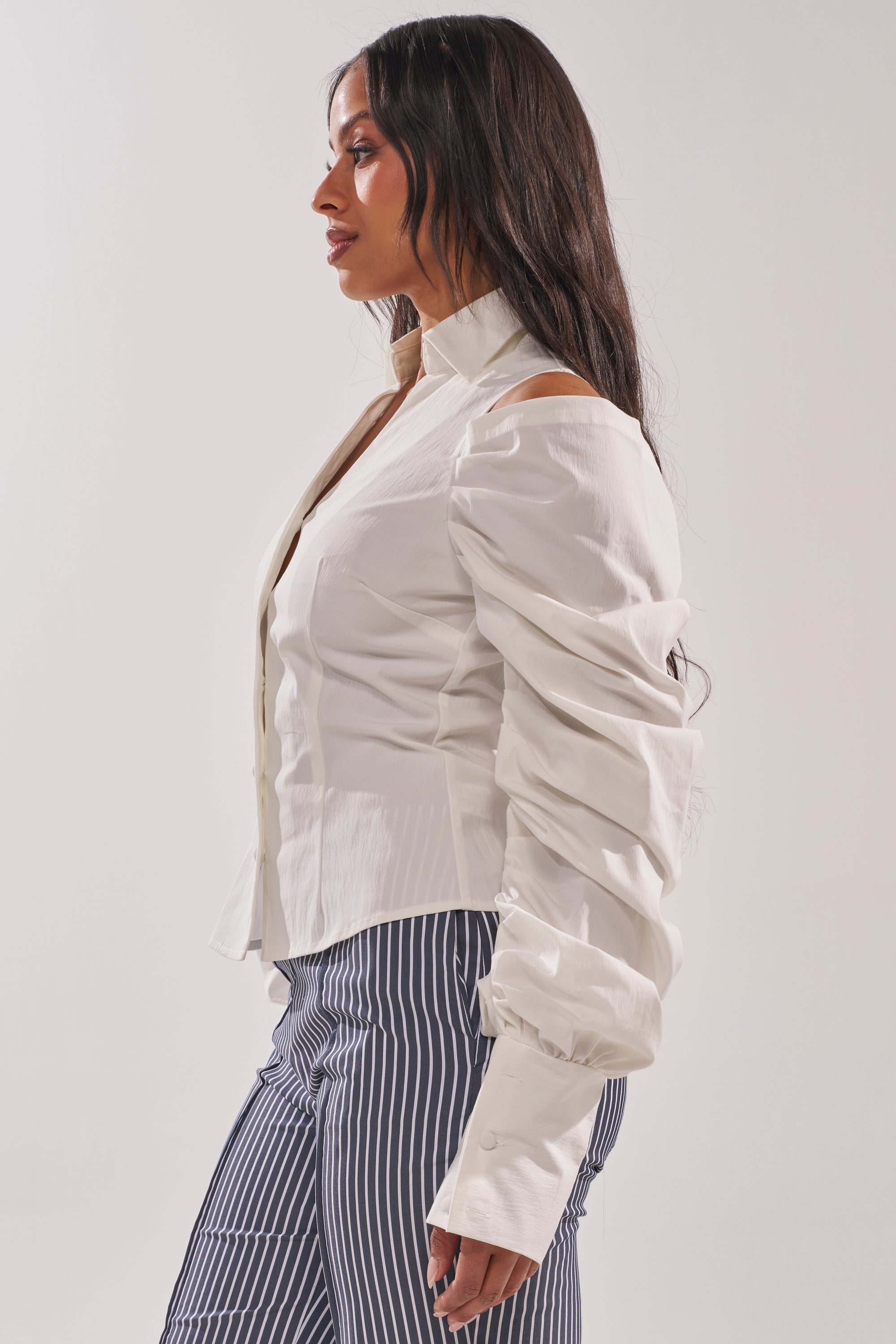 A woman stands in profile wearing the CRAZY IN LOVE WHITE BUTTON DOWN BLOUSE with ruched sleeves and blue-and-white vertically striped pants against a plain light background. 3