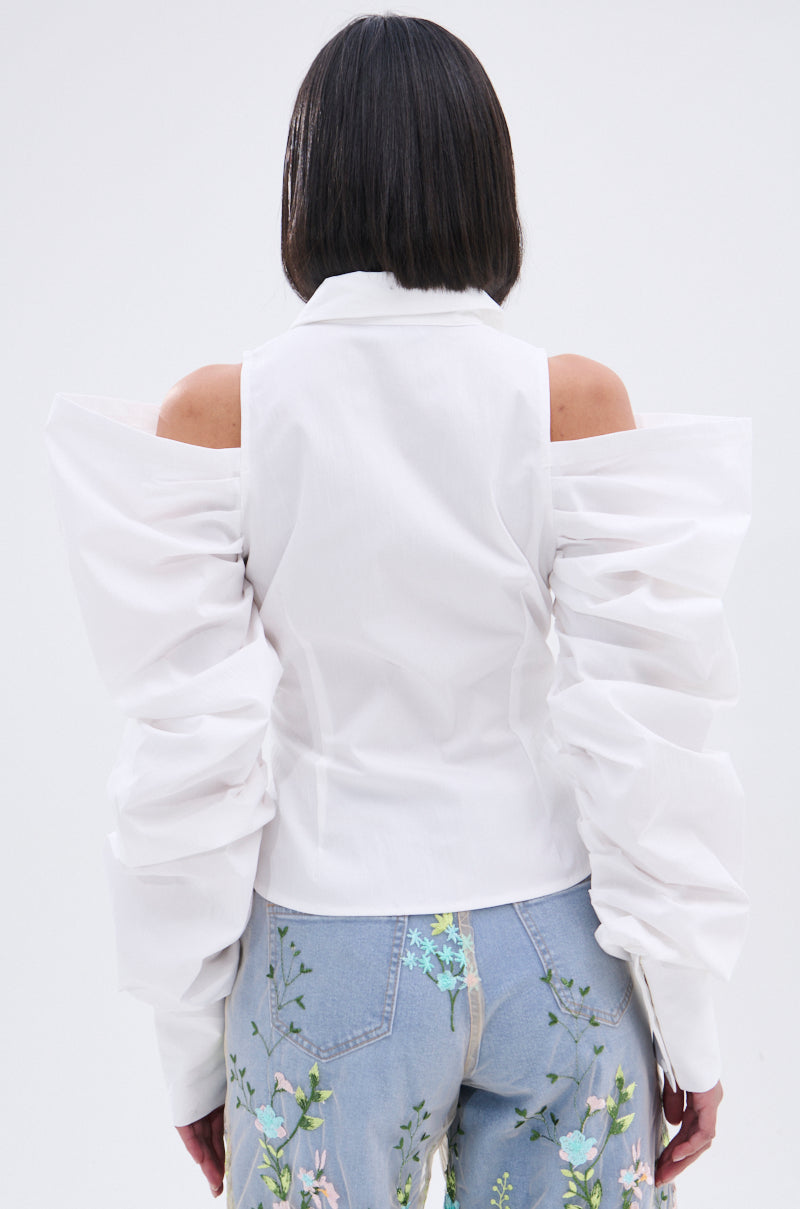 A person with a bob haircut is seen from behind wearing the CRAZY IN LOVE BUTTON DOWN BLOUSE featuring voluminous, ruched statement sleeves and a cold shoulder design, paired with light blue floral embroidered jeans.