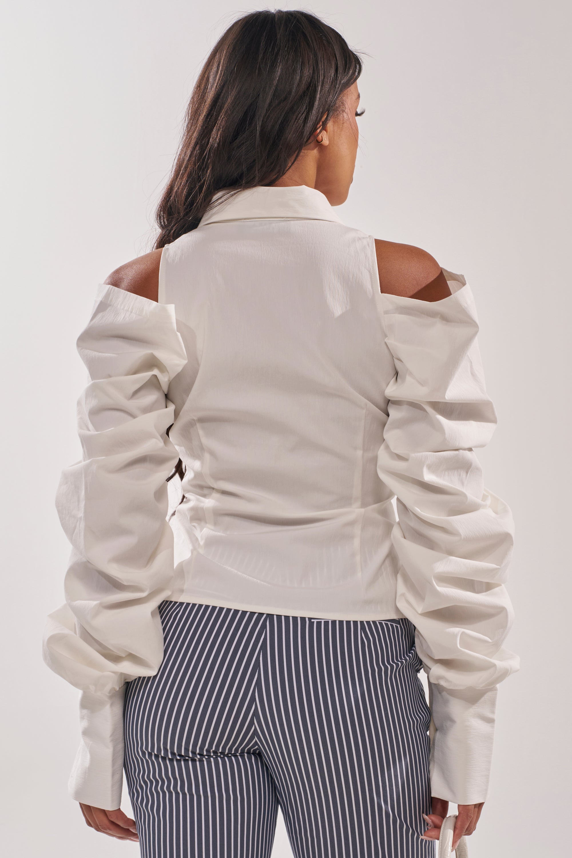 A woman faces away, wearing the CRAZY IN LOVE WHITE BUTTON DOWN BLOUSE with voluminous ruched sleeves and high-waisted black-and-white striped pants, set against a plain light background. 4