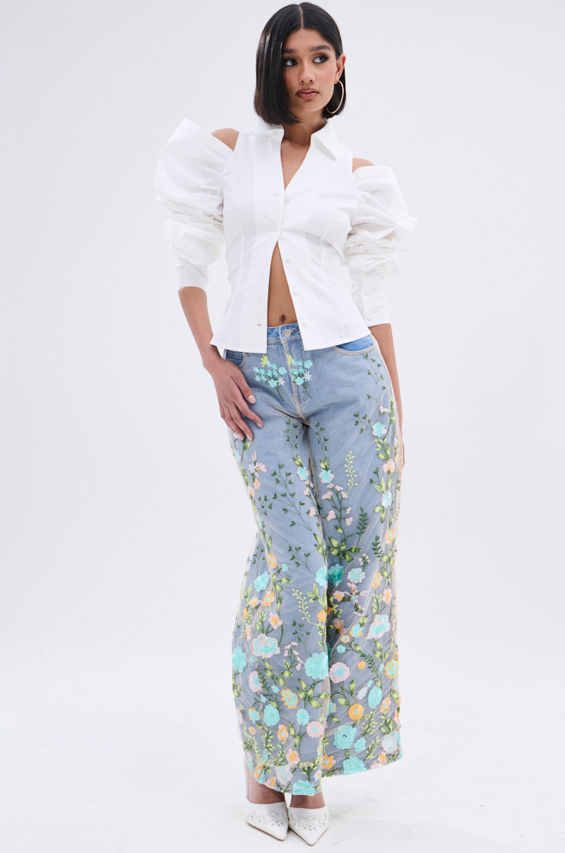 A woman wears the CRAZY IN LOVE BUTTON DOWN BLOUSE in white with statement sleeves and high-waisted, wide-leg blue jeans featuring a colorful floral pattern, standing against a plain white background.