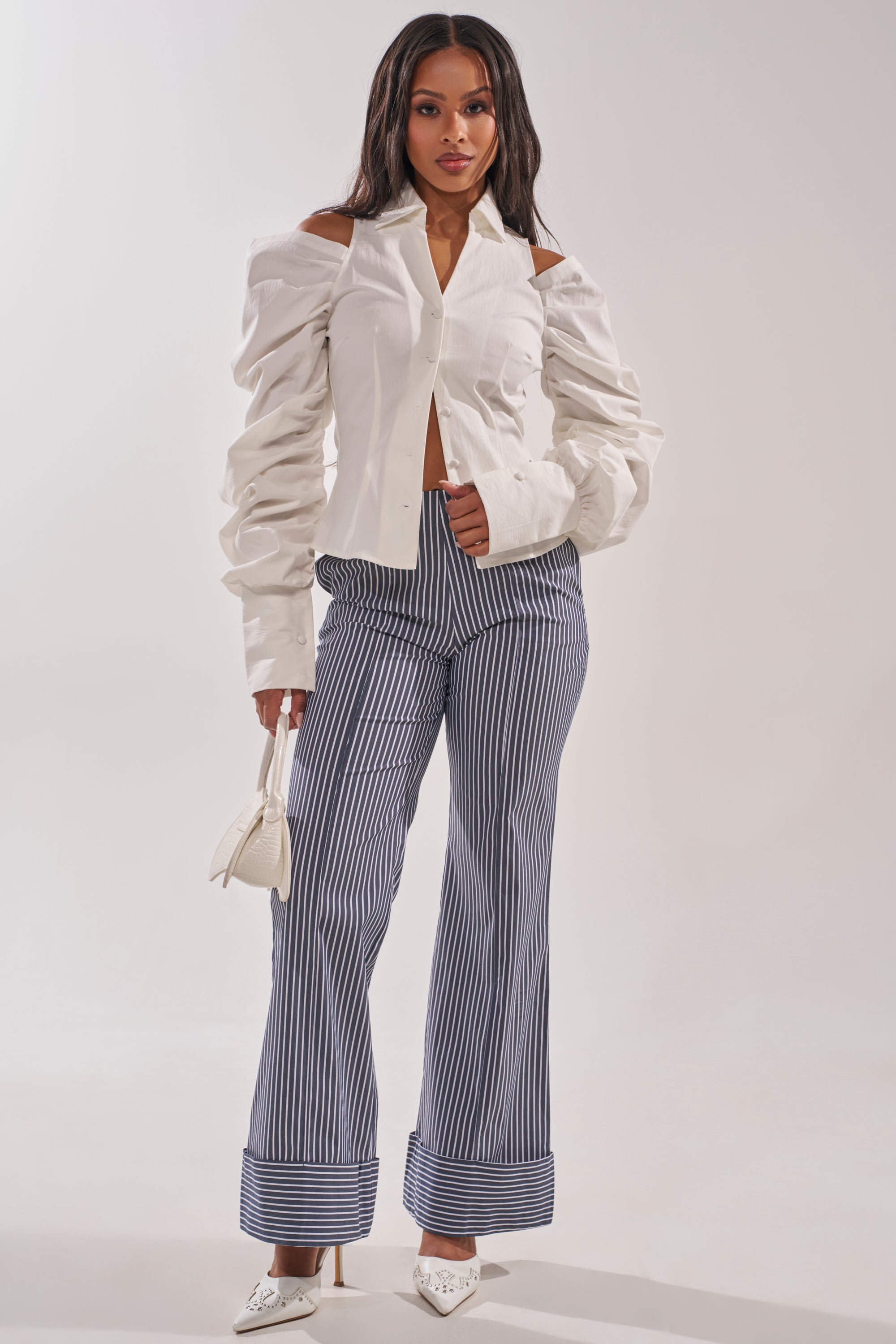 A woman stands against a light background wearing the CRAZY IN LOVE WHITE BUTTON DOWN BLOUSE with puffed sleeves, striped wide-leg trousers, white pointed heels, and holding a small white handbag. 5