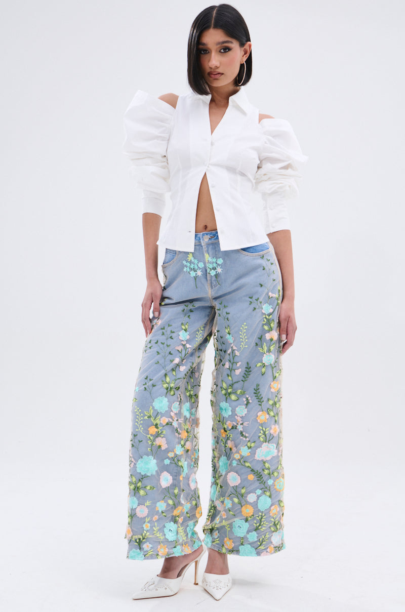 A woman wears the CRAZY IN LOVE BUTTON DOWN BLOUSE with statement sleeves, paired with high-waisted light blue, wide-leg pants with floral embroidery, white heels, and a sleek straight hairstyle.