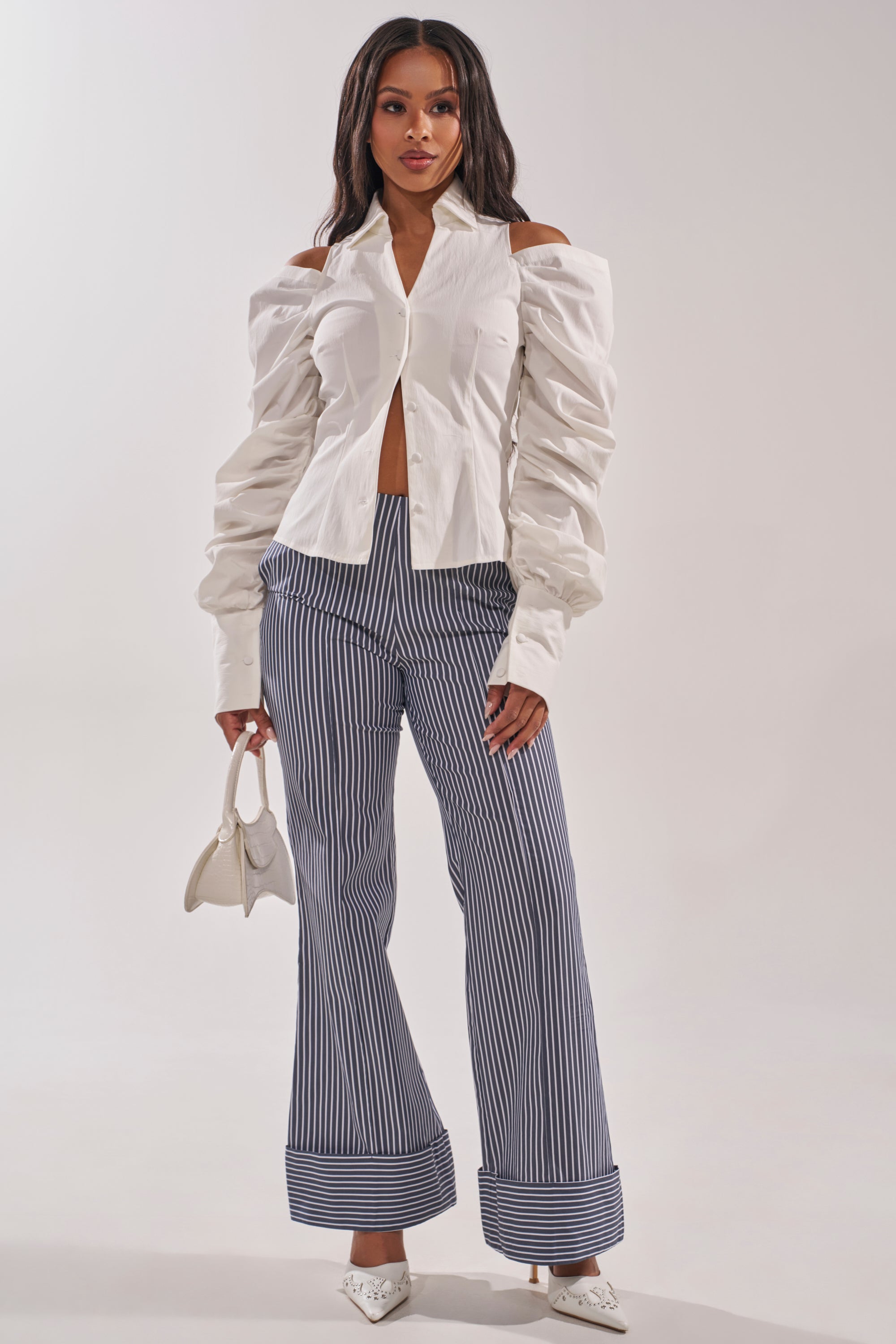 A woman wears the CRAZY IN LOVE WHITE BUTTON DOWN BLOUSE with ruched sleeves, paired with blue and white striped wide-leg pants, white pointed heels, and a small white handbag against a plain background. 6