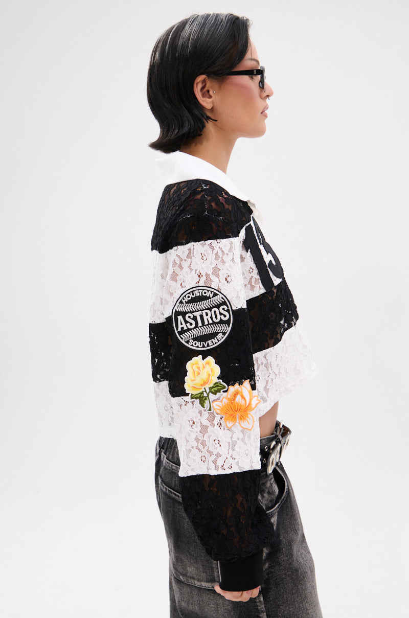 A person in sunglasses, faded black jeans, and the GIVE ME TIME LACE JERSEY—a black and white varsity-inspired crop top with an "ASTROS" patch and yellow flowers—poses in profile against a plain white background. 3