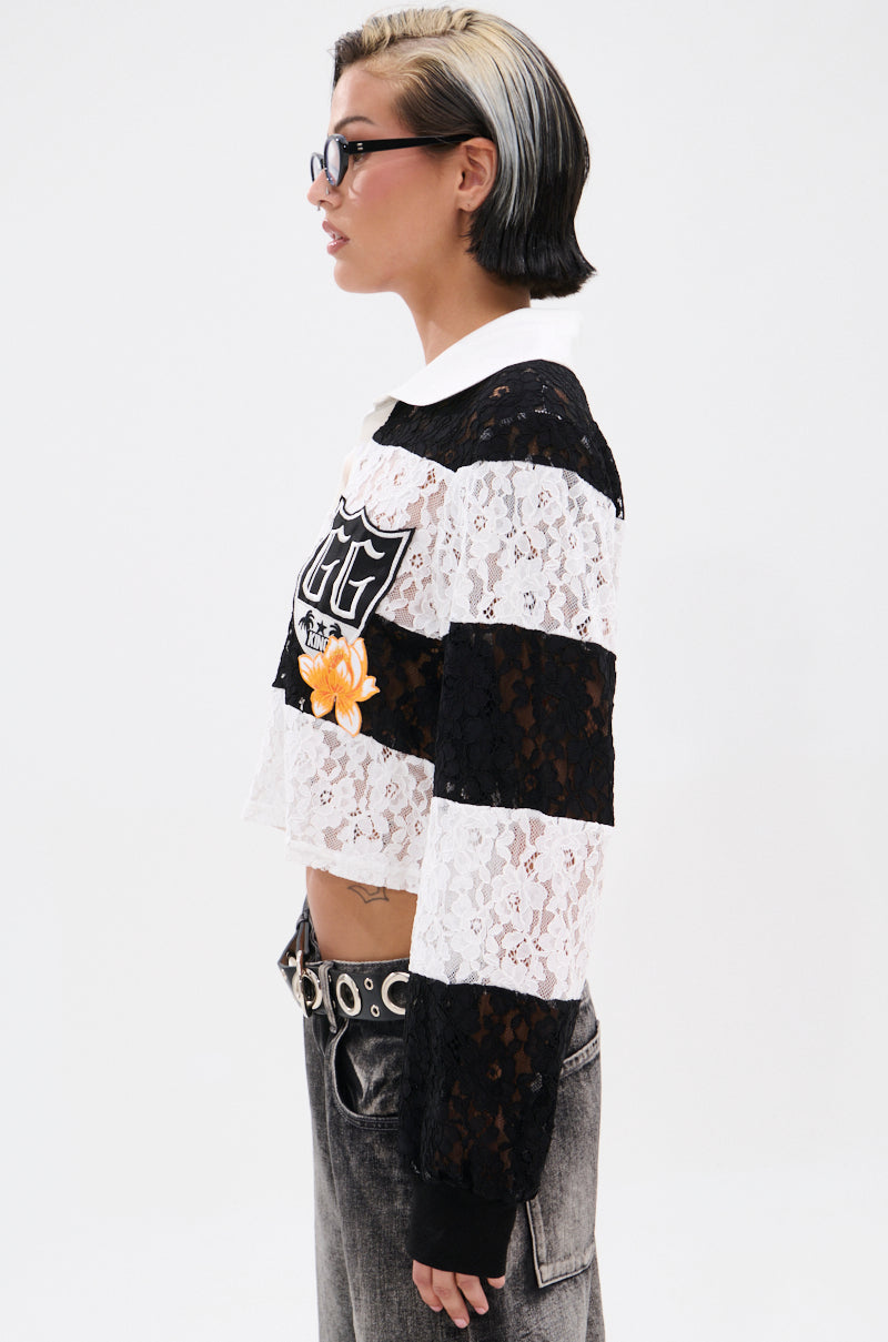 A person with short black and blonde hair, wearing glasses, models the GIVE ME TIME LACE JERSEY—a varsity-style black-and-white striped long-sleeve shirt with lace details and embroidery—paired with gray jeans and a studded belt, side view. 4