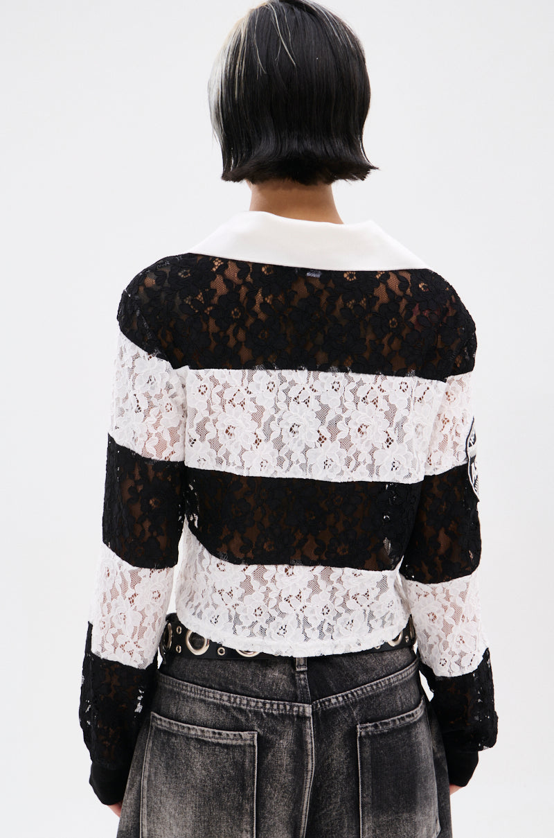 A person with short, dark hair and a white streak stands facing away, wearing the GIVE ME TIME LACE JERSEY—a sheer, varsity-inspired black and white striped lace top with a white collar—paired with faded black jeans featuring silver grommet details. 5
