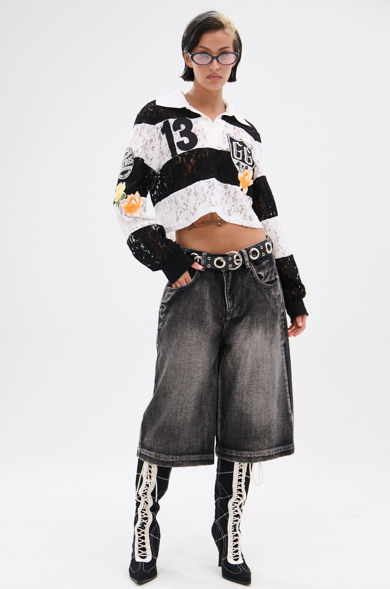 A person stands confidently in the GIVE ME TIME LACE JERSEY, styled with bold black and white stripes, oversized faded knee-length shorts, lace-up high-heeled boots, a studded belt, and rectangular glasses. 6