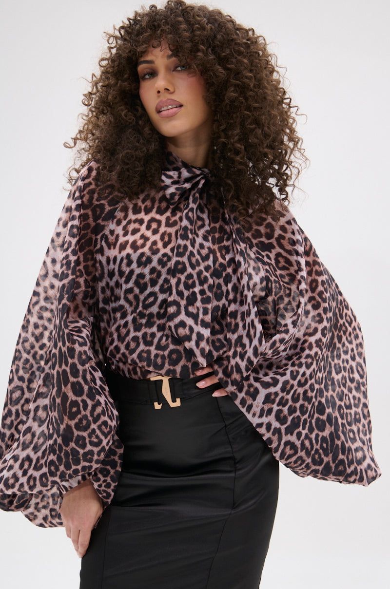 A woman with curly hair poses confidently in the ROXY CHEETAH BLOUSE—a sheer, long-sleeved chiffon top with a bow at the neck—paired with a high-waisted black skirt against a plain background.
