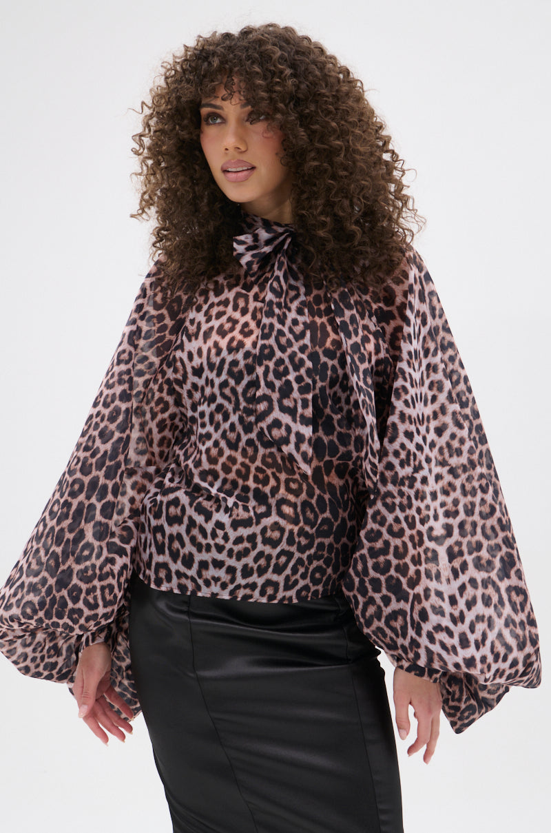 A woman with curly hair models the ROXY CHEETAH BLOUSE—sheer chiffon with voluminous sleeves and a neck bow—paired with a fitted black leather skirt against a white background, making this blouse a true statement piece.