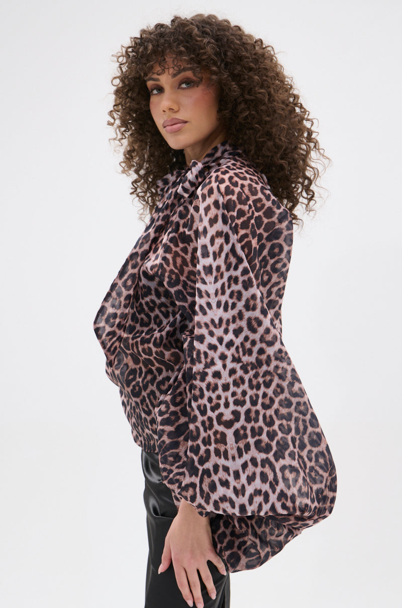 A woman with curly hair poses sideways against a white background, making a bold statement in the ROXY CHEETAH BLOUSE—a sheer chiffon top with long sleeves—paired with black pants.