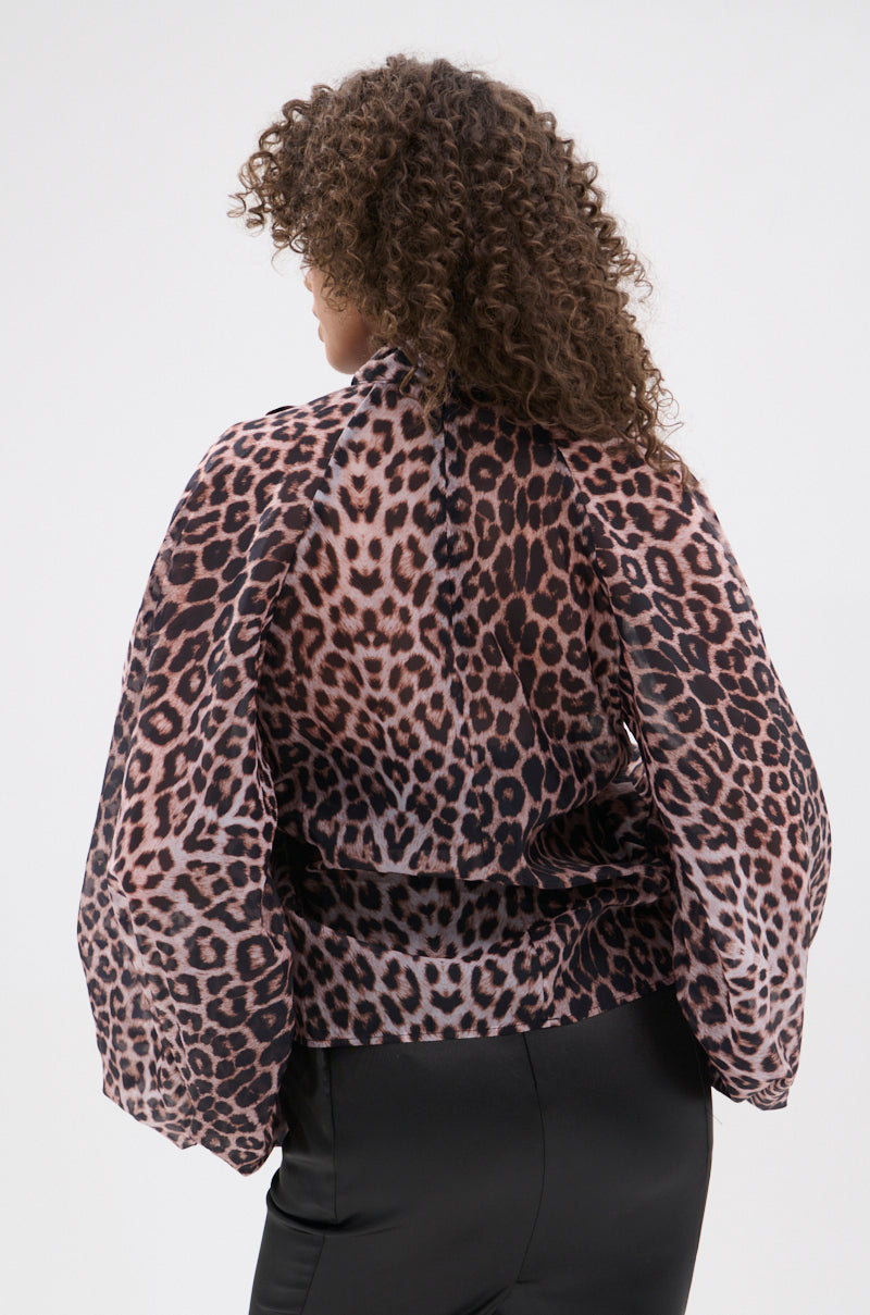 A person with curly hair is shown from the back, wearing the ROXY CHEETAH BLOUSE—a sheer chiffon, long-sleeved top in pink and brown cheetah print—paired with black pants against a plain white background.