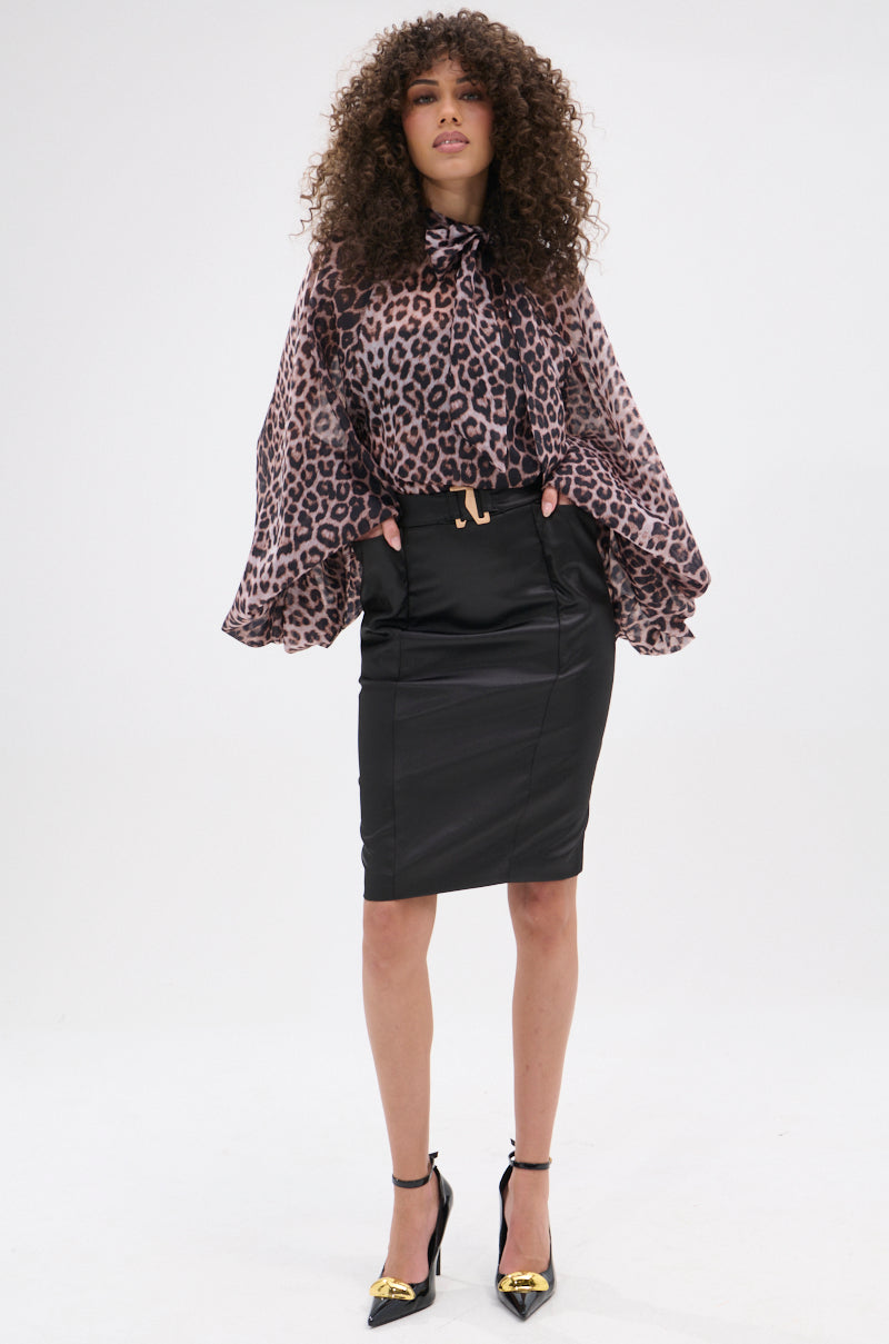 A woman with curly hair confidently wears the ROXY CHEETAH BLOUSE, tucked into a fitted black pencil skirt and paired with black heels featuring gold detail. She stands with hands in her pockets against a plain white background.