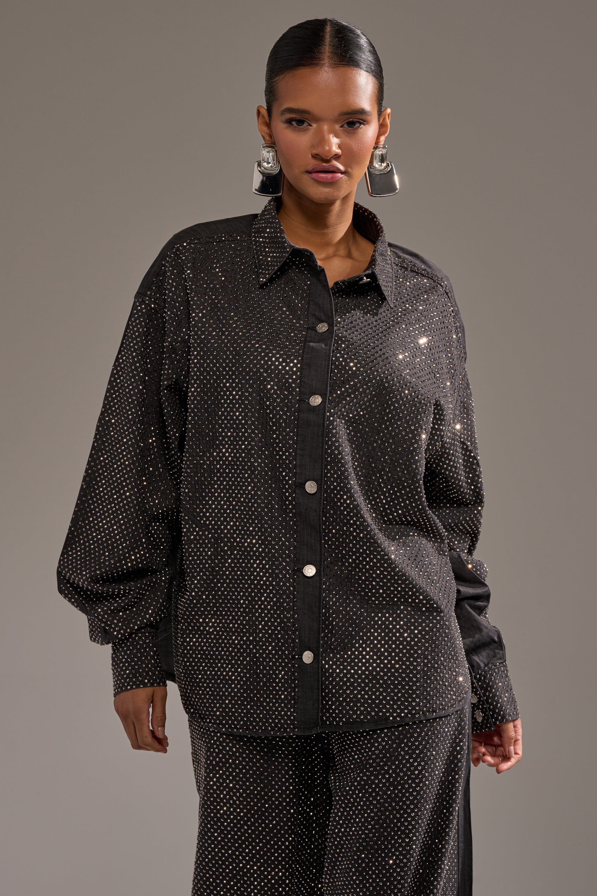 A woman with slicked-back hair wears the BLING BLING DENIM BUTTON DOWN TOP IN BLACK, covered in sparkling metallic dots, paired with matching pants and accessorized with large geometric silver earrings. 1