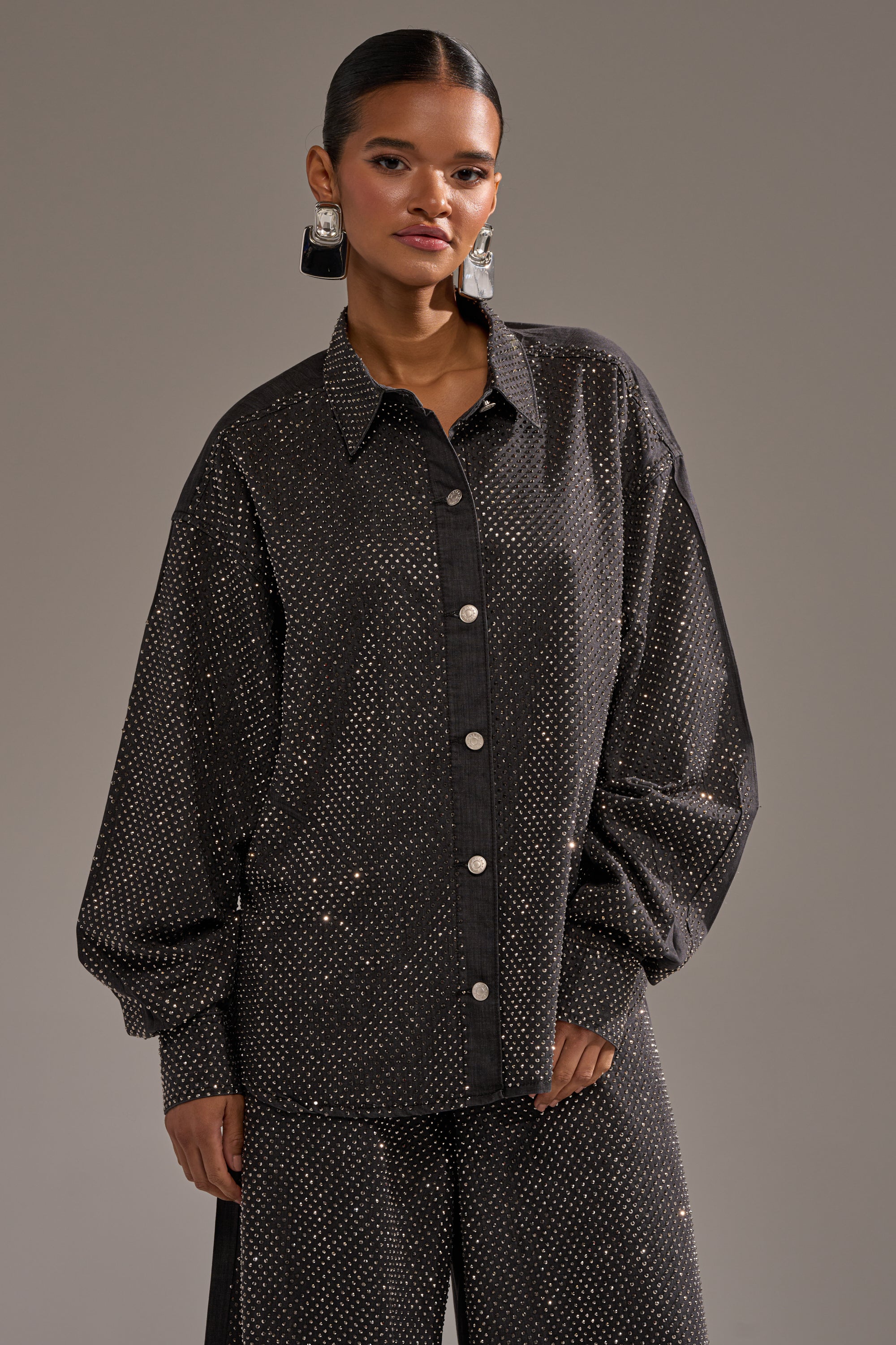 A woman wears the BLING BLING DENIM BUTTON DOWN TOP IN BLACK, a loose-fitting shirt with a small shimmering dot pattern, paired with matching wide-leg pants. Her hair is pulled back, and she accessorizes with geometric earrings against a plain background. 2