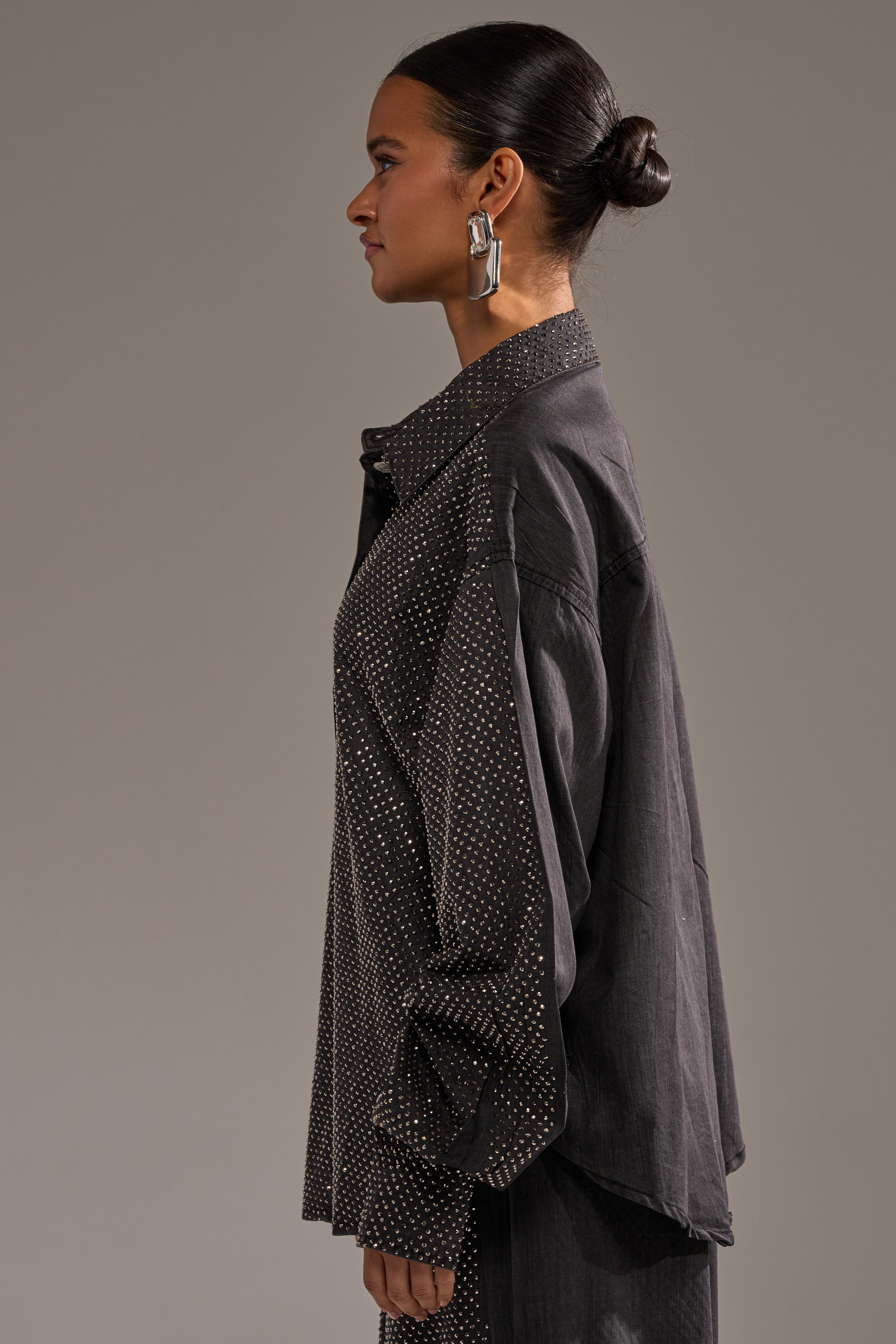 A woman with her hair in a bun, wearing large rectangular earrings and the BLING BLING DENIM BUTTON DOWN TOP IN BLACK with a dotted pattern, stands in profile against a plain background. 3