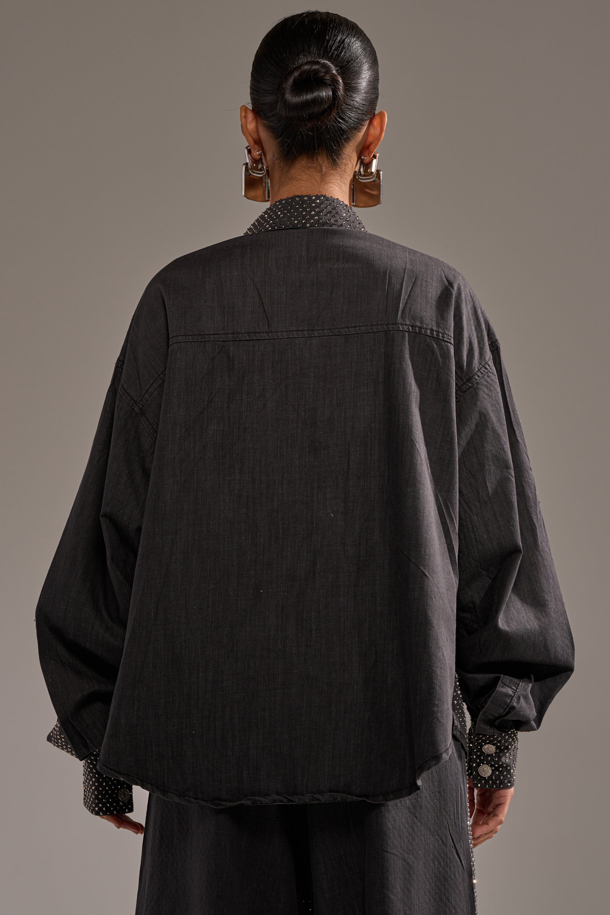 A person with dark hair in a neat bun, wearing large hoop earrings and the BLING BLING DENIM BUTTON DOWN TOP IN BLACK, is shown from the back against a plain gray background. 4