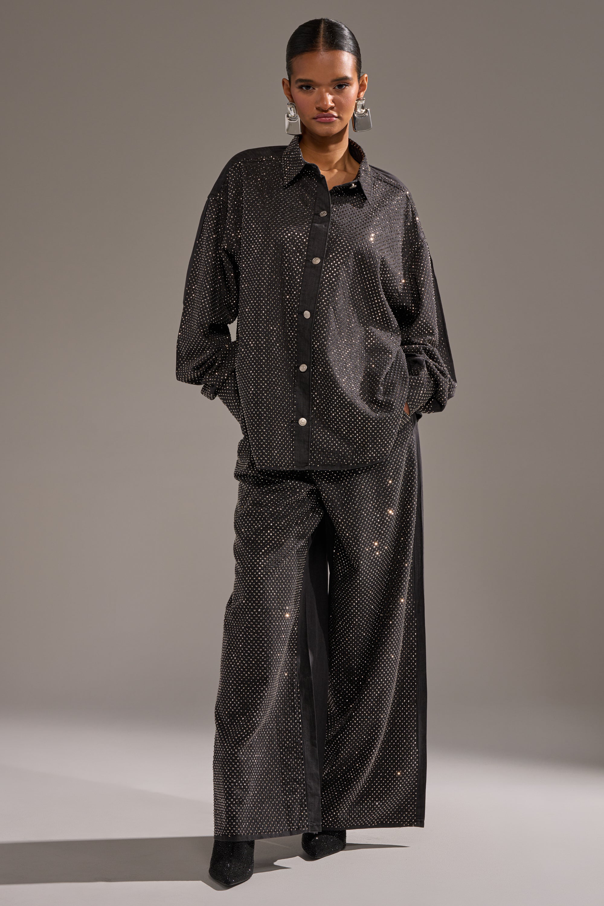 A woman stands confidently in the BLING BLING DENIM BUTTON DOWN TOP IN BLACK—an oversized black denim top with a subtle shimmering dot pattern—paired with matching wide-leg pants, hands in pockets, against a plain gray background. 5