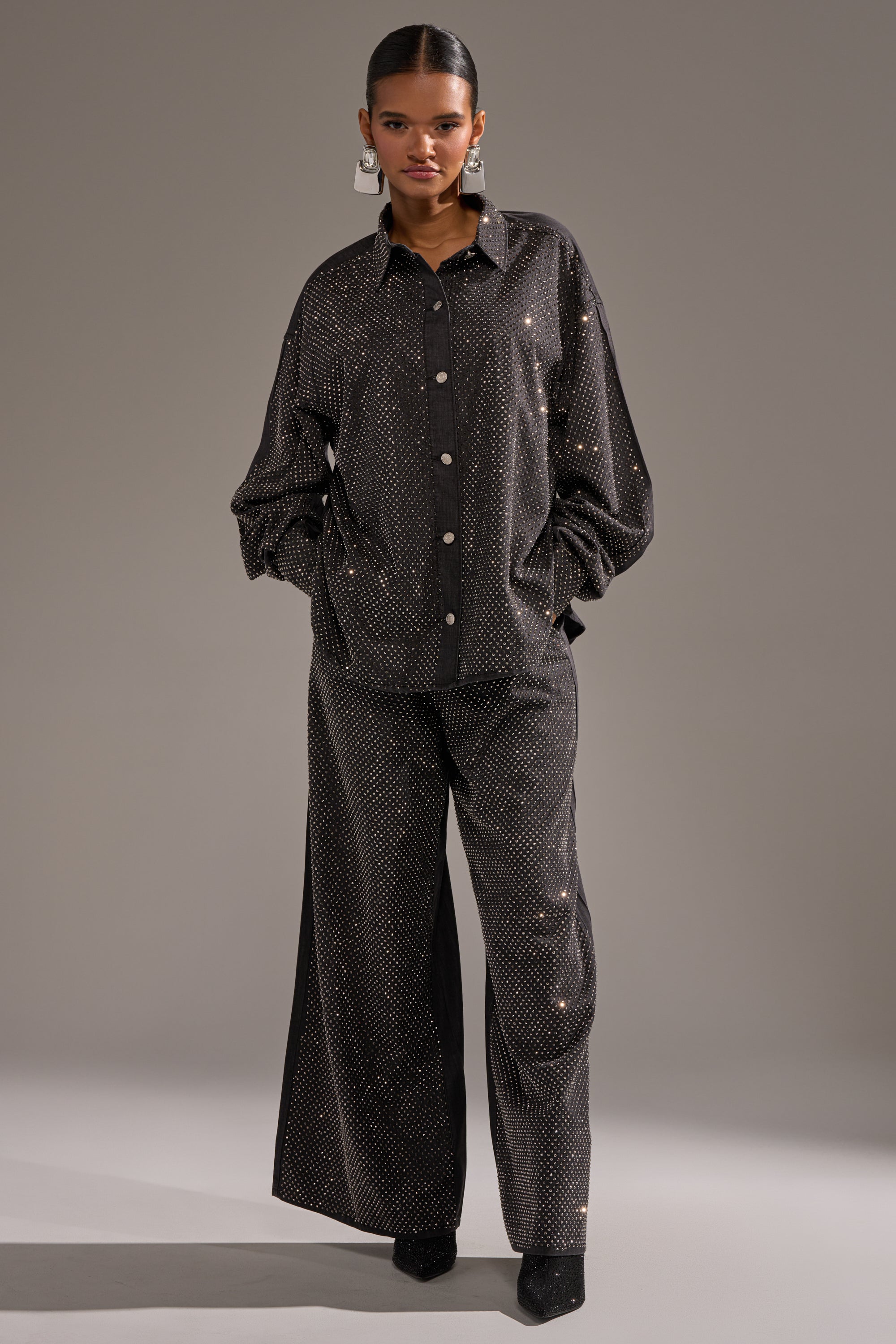 A person stands against a neutral background wearing the BLING BLING DENIM BUTTON DOWN TOP IN BLACK, paired with wide-leg pants featuring a subtle sparkling dotted pattern, black shoes, and large hoop earrings. 6