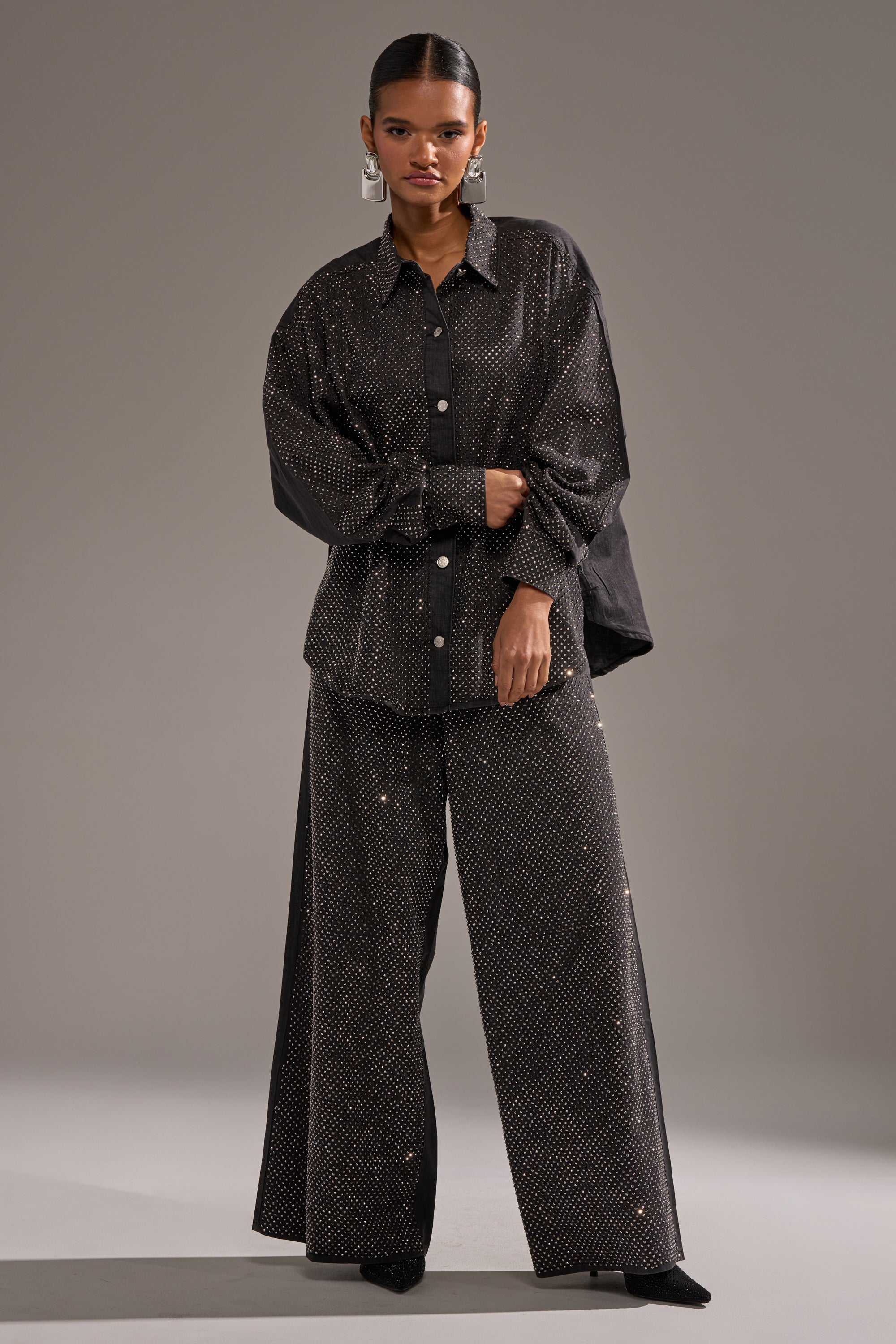 A woman wears the BLING BLING DENIM BUTTON DOWN TOP IN BLACK, a relaxed and oversized piece. She pairs it with wide-leg polka dot pants, accessorizes with statement earrings, and poses confidently against a plain background. 7