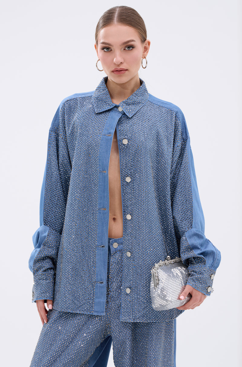 A woman with slicked-back hair and hoop earrings wears the BLING BLING DENIM BUTTON DOWN TOP with matching pants, holding a metallic silver purse. She stands confidently against a plain light background.