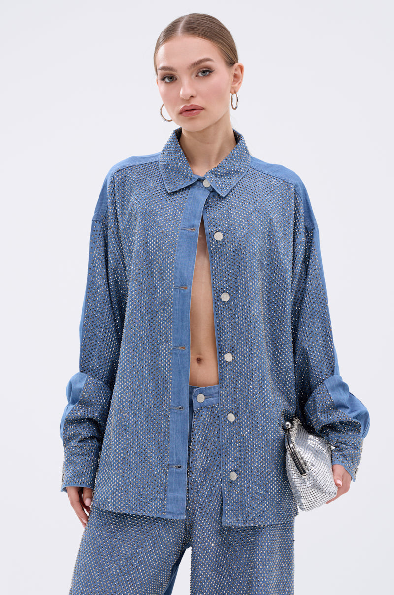 A woman with slicked-back hair poses against a white background, wearing the BLING BLING DENIM BUTTON DOWN TOP left mostly unbuttoned with matching pants. She accessorizes with a small silver purse and large hoop earrings.