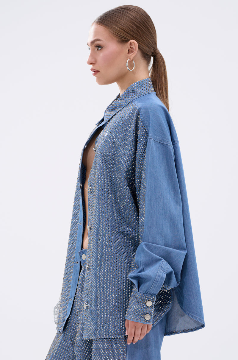 A woman with straight light brown hair in a low ponytail wears the BLING BLING DENIM BUTTON DOWN TOP adorned with rhinestones, paired with matching wide-legged pants. She stands in profile against a plain light background.
