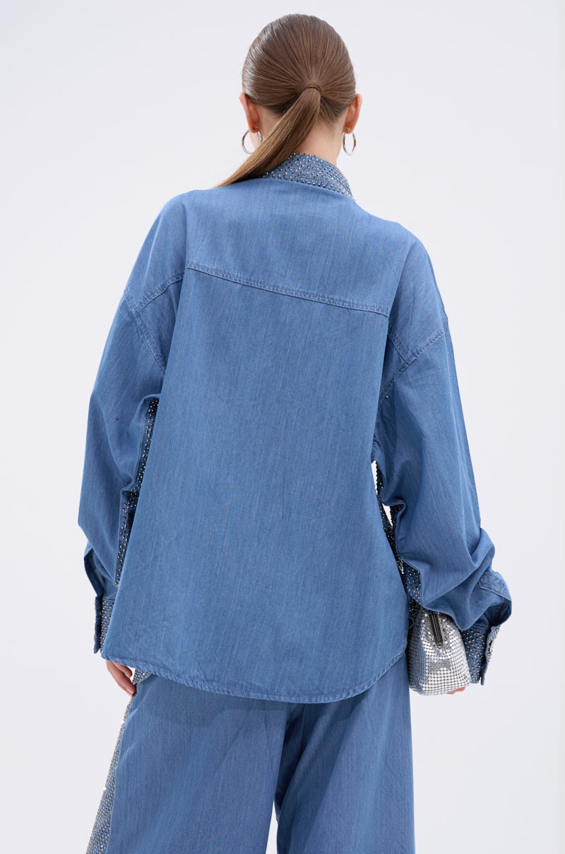 A person with long hair tied back is seen from behind, wearing an oversized BLING BLING DENIM BUTTON DOWN TOP and wide-leg jeans. A shiny silver bag adds a touch of sparkle to the relaxed, casual look.