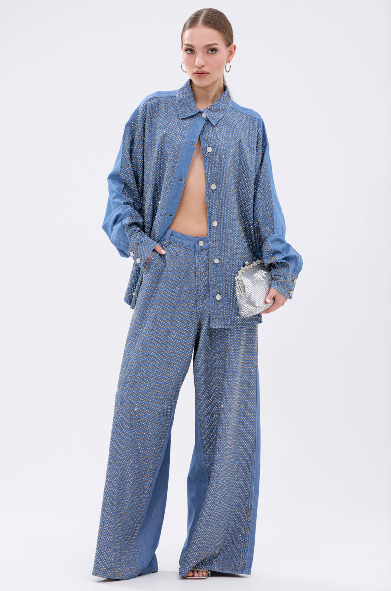 A woman models the BLING BLING DENIM BUTTON DOWN TOP in light blue, styled open with matching wide-leg pants. She accessorizes with a silver handbag, hoop earrings, and has her hair pulled back.