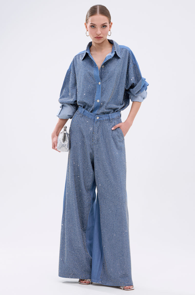 A woman poses in the oversized, sparkling BLING BLING DENIM BUTTON DOWN TOP with matching wide-leg pants, styled with silver heels, hoop earrings, and a small bag. A white background highlights her shimmering look.