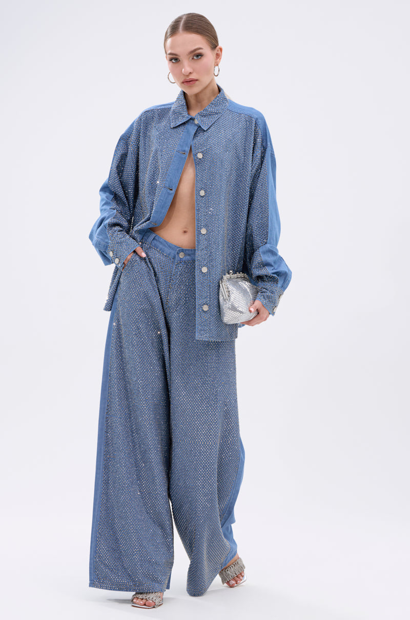 A woman poses against a plain background wearing the BLING BLING DENIM BUTTON DOWN TOP—an oversized, sparkly blue shirt with rhinestones—styled unbuttoned to show her midriff, paired with matching wide-leg pants, silver heels, and a small silver clutch.