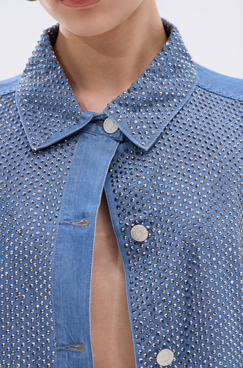 A person wears the BLING BLING DENIM BUTTON DOWN TOP, a light blue denim jacket featuring small silver studs, rhinestones, and metallic buttons. The jacket is partially unbuttoned to reveal some skin underneath.